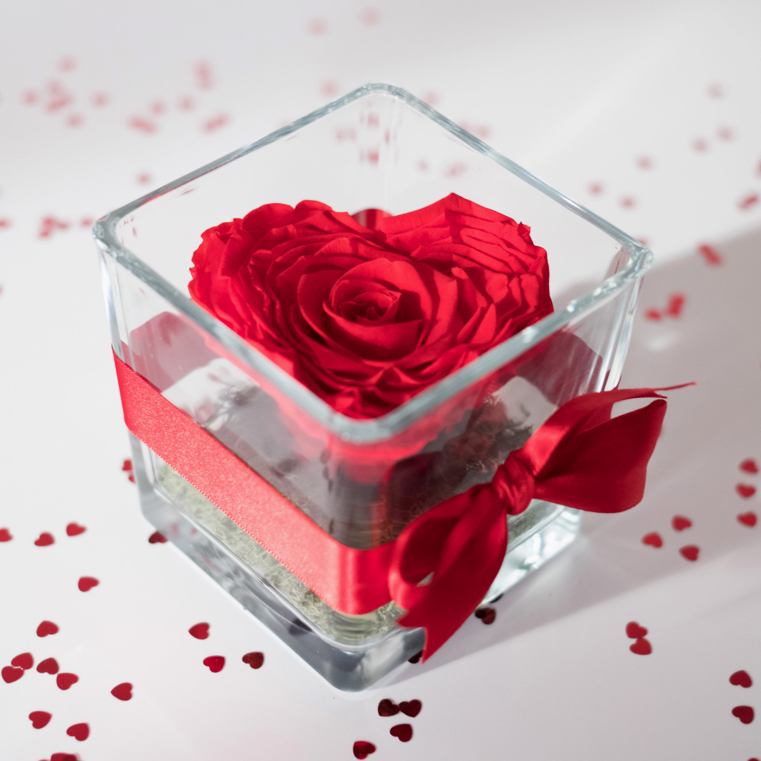rosa rossa a forma di cuore stabilizzata nel contenitore di vetro con nastro rosso su tavolo bianco