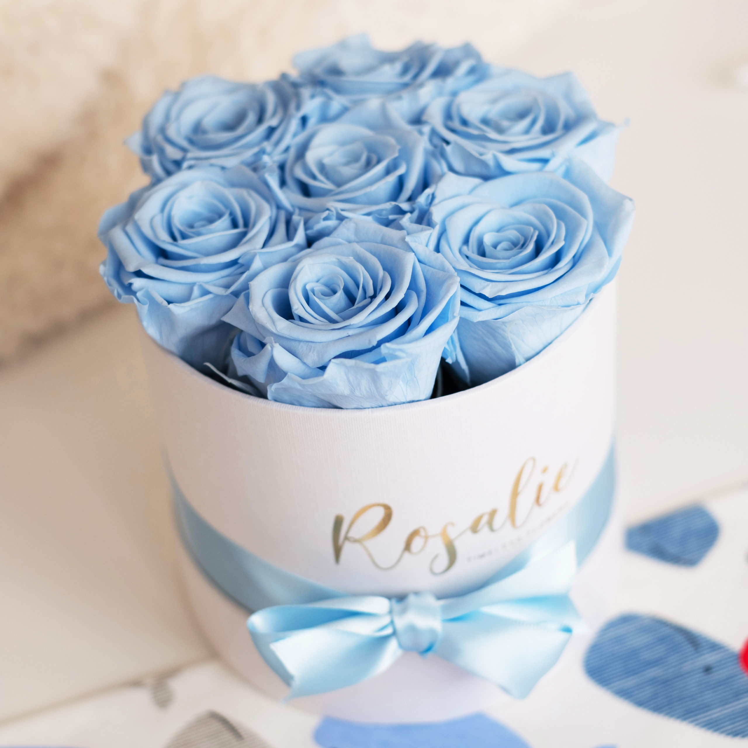 scatola tonda con 7 rose stabilizzate di color azzurro e nastro di raso azzurro attorno. Su tavolo bianco. Vista dall'alto.