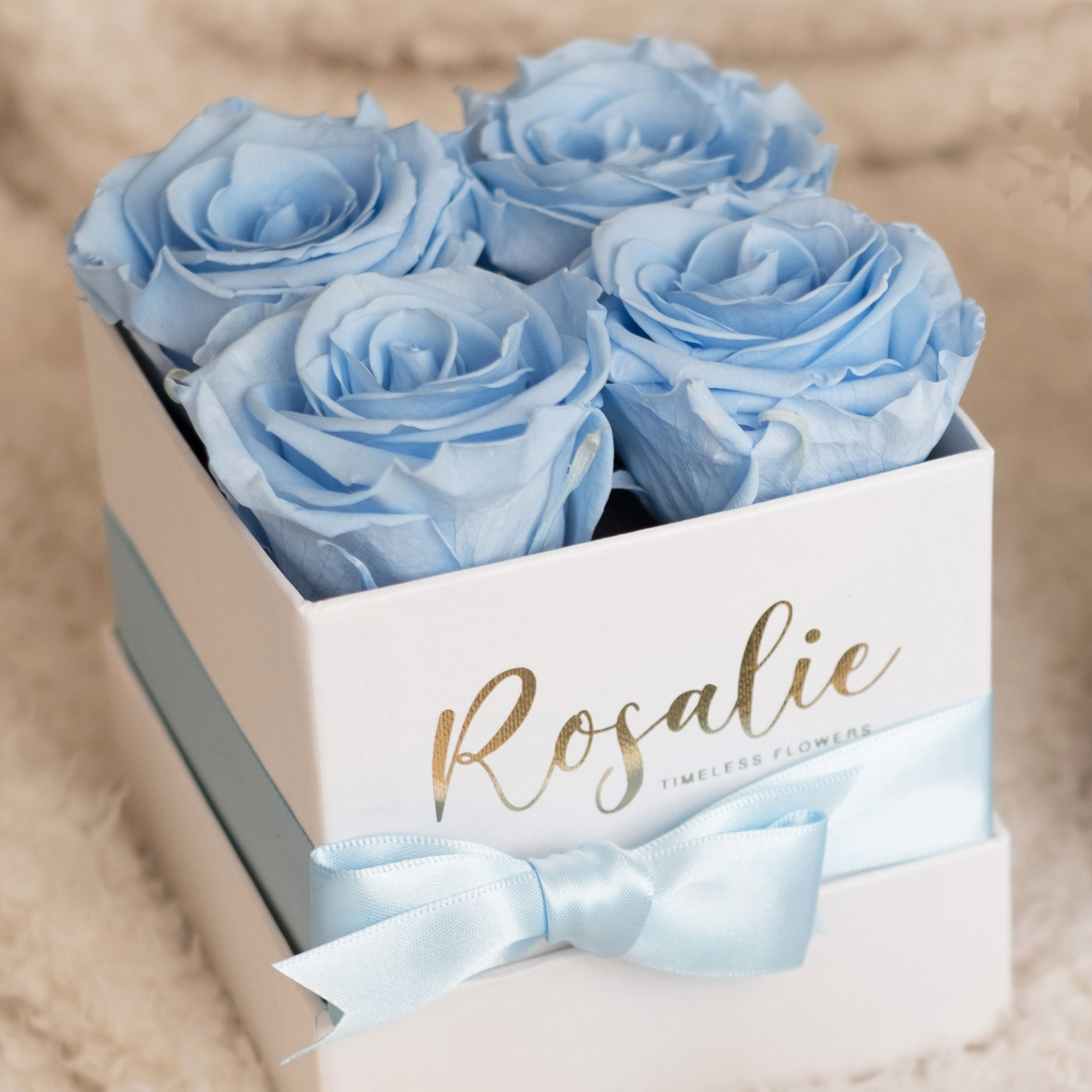 scatola bianca quadrata con 4 rose stabilizzate azzurre e nastro azzurro, poggiata su coperta bianca.