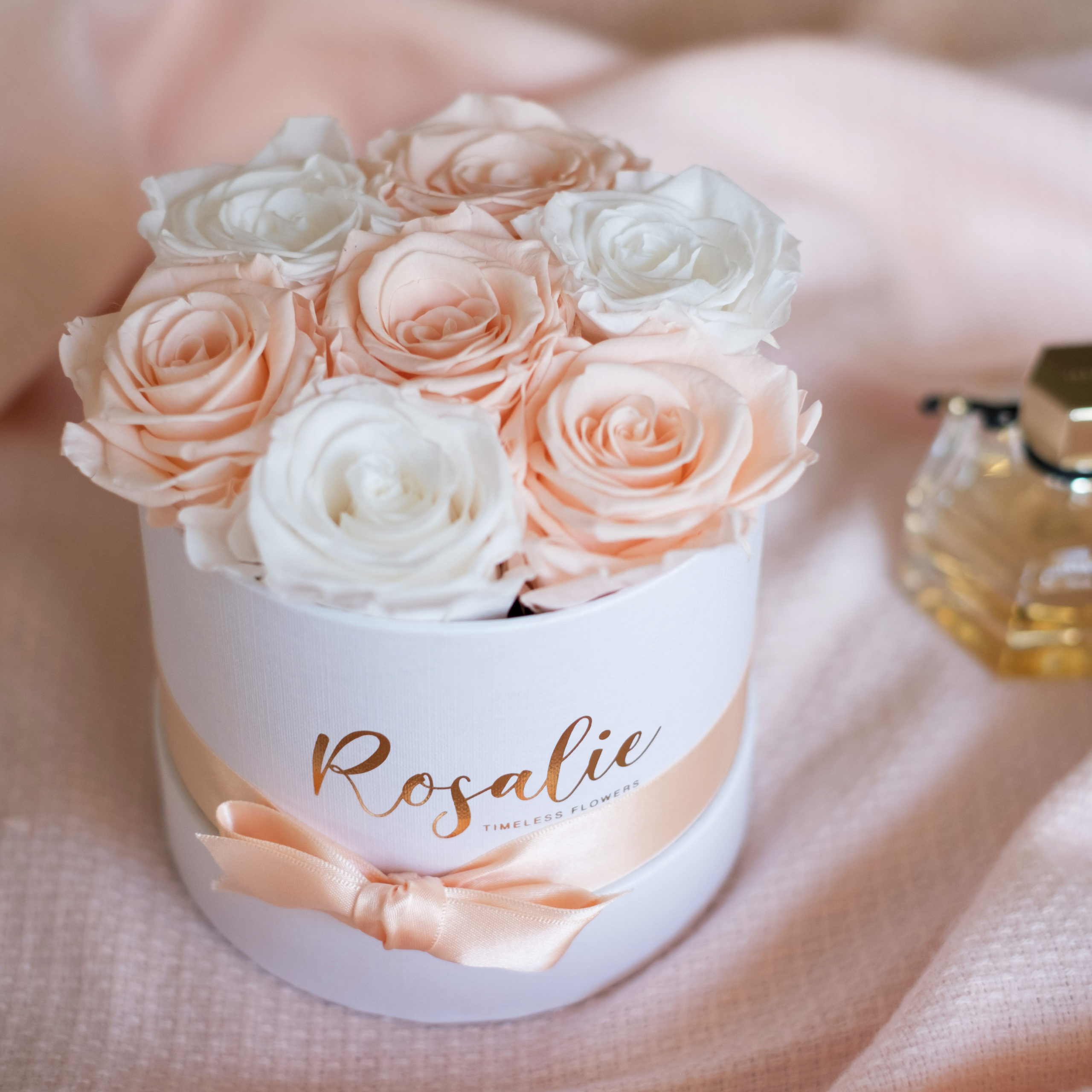 scatola tonda con 7 rose stabilizzate di cui 4 rosa porcellena e 3 bianche su tessuto rosa accanto ad un profumo