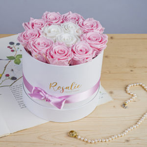 scatola da 12 rose stabilizzate di cui 9 di color rosa e 3 centrali bianche. Con nastro rosa attorno. Appoggiata su libro con accanto collana di perle. Su tavolo di legno chiaro.