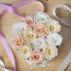 scatola box tonda da 12 rose stabilizzate visto dall'alto. Rose di color rosa porcellana, bianco e panna. Nastro rosa e bianco vicino alla scatola. Tutto appoggiato su tavolo di legno chiaro.