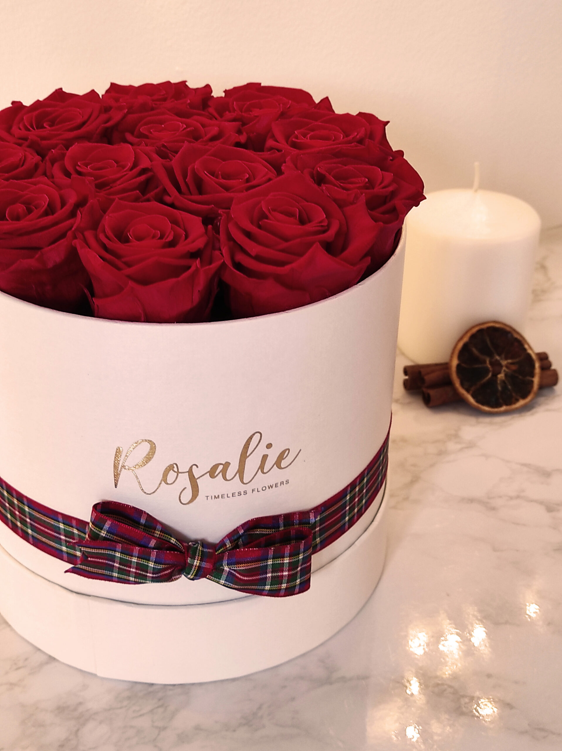 scatola tonda bianca con 12 rose rosse stabilizzate e nastro natalizio su tavolo di marmo con candela