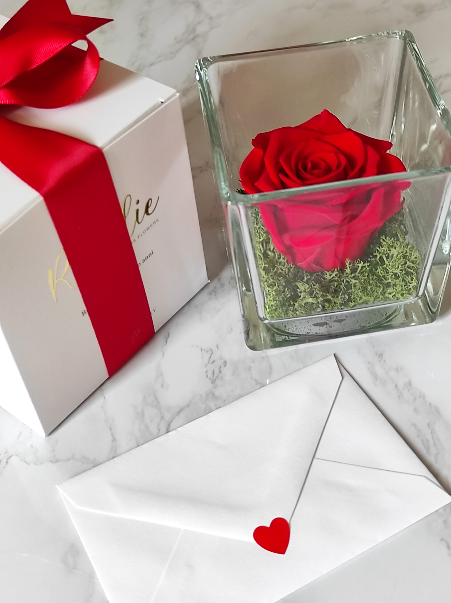 rosa rossa stabilizzata in cubo di vetro accanto a confezione regalo con nastro rosso e biglietto